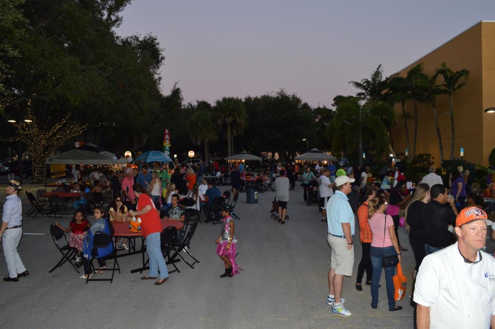 People stand and sit in chairs at tables in area of street near vendors